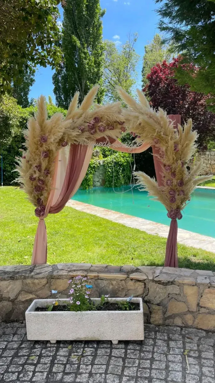 Un arco nupcial decorado con hierba de la pampa y tela rosa pálido se alza en un patio con vistas a una piscina y un jardín.