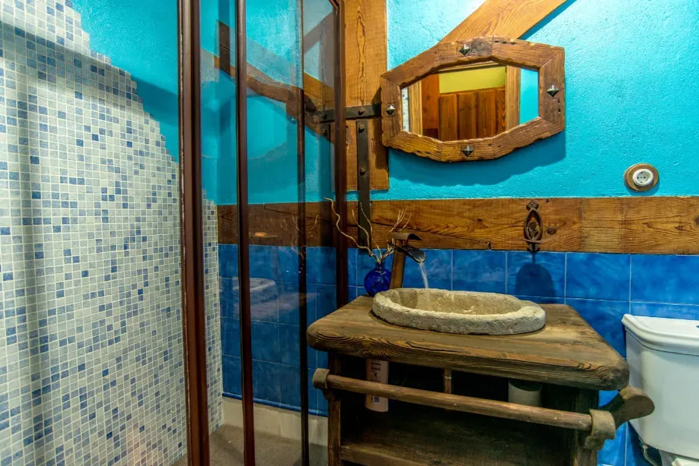 Baño rústico con lavabo de piedra, tocador de madera, ducha de azulejos y espejo decorativo sobre paredes de color azul brillante.