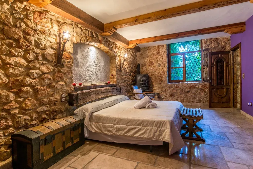 Un dormitorio rústico con paredes de piedra, una cama doble con ropa de cama blanca, vigas de madera, un baúl de madera y una ventana.