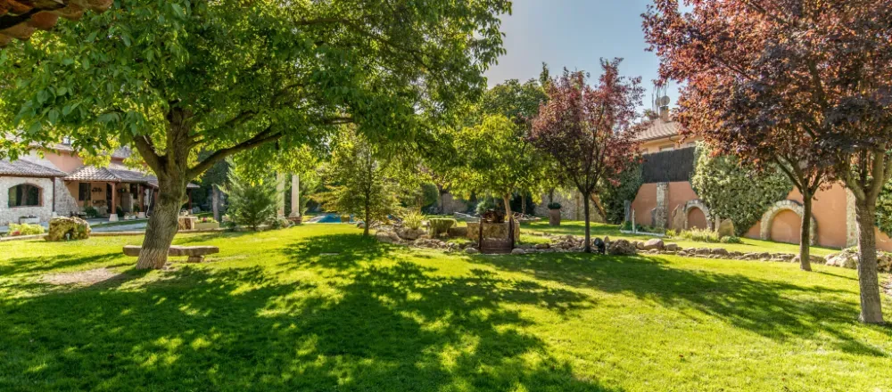 Un jardín exuberante y soleado con amplios céspedes verdes, árboles maduros y edificios rústicos de piedra bajo un cielo azul despejado.