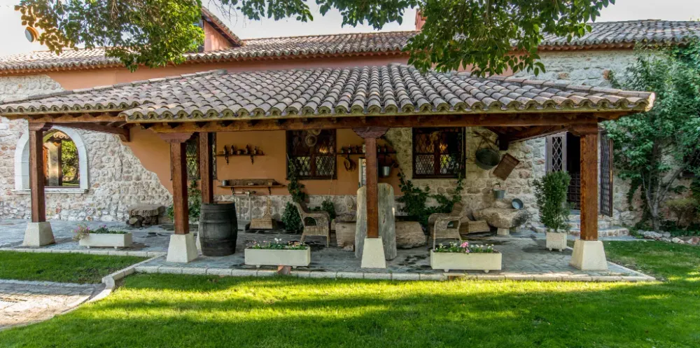Un patio cubierto con vigas de madera y techo de tejas, adosado a una rústica casa de campo de piedra con un césped delante.