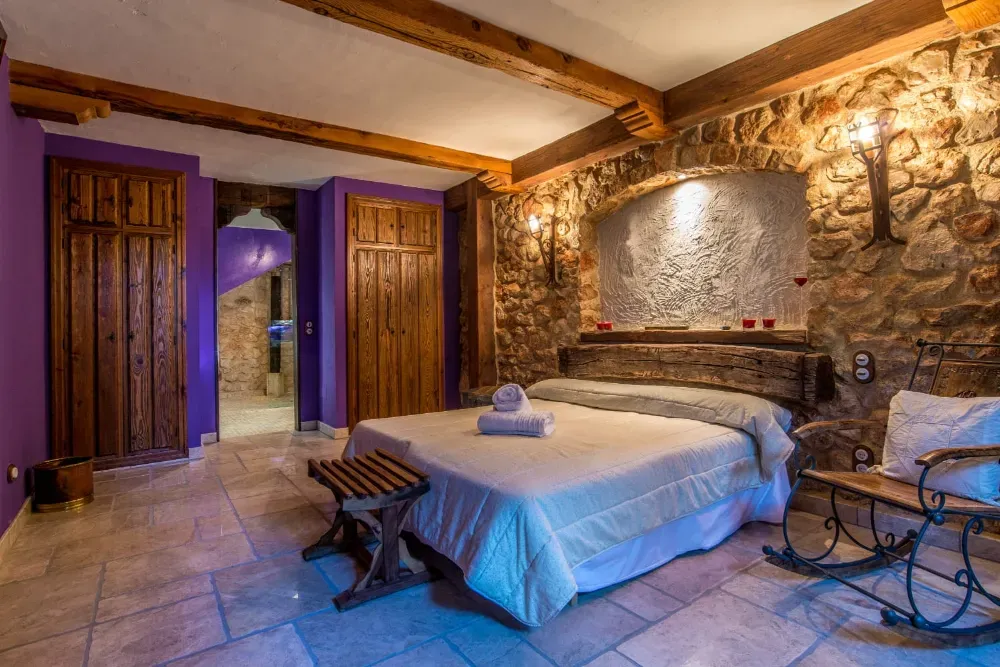 Un dormitorio rústico, iluminado con velas, con una pared de piedra, vigas de madera, detalles en color púrpura, una cama y una mecedora sobre suelo de baldosas.