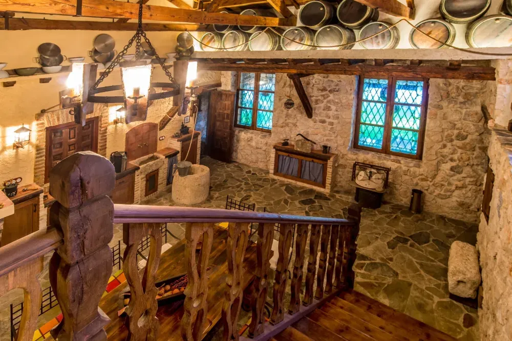 Interior rústico con paredes de piedra, escalera de madera, barriles de vino suspendidos y una lámpara de araña en una habitación con suelo de piedra.