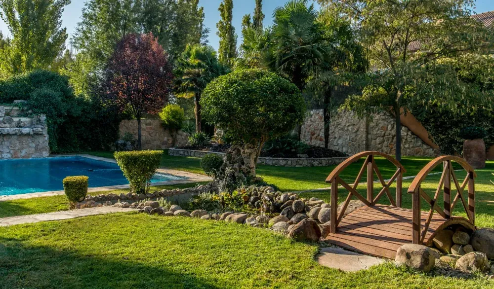 Un jardín exuberante con una piscina azul, un pequeño puente de madera, setos bien cuidados y árboles maduros bajo un cielo azul.