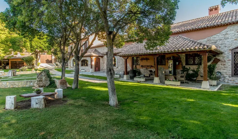 En un día soleado, un edificio de piedra con tejado de tejas y un patio cubierto se alza junto a un frondoso césped verde y árboles.
