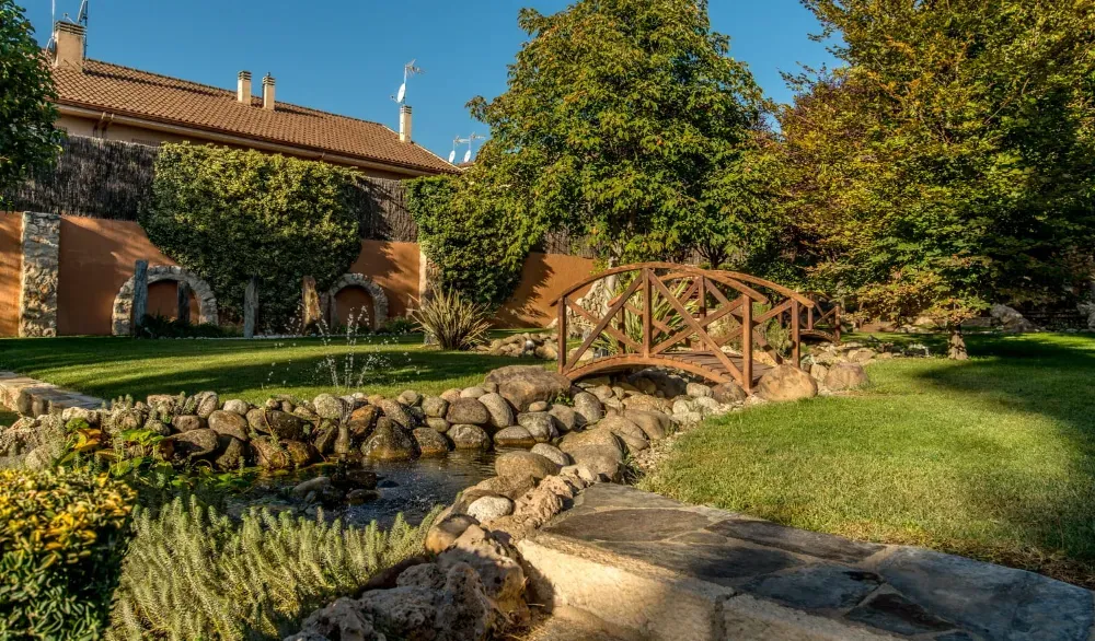 Un puente de madera arqueado sobre un arroyo bordeado de piedras en un jardín frondoso y soleado, con una casa de piedra al fondo.