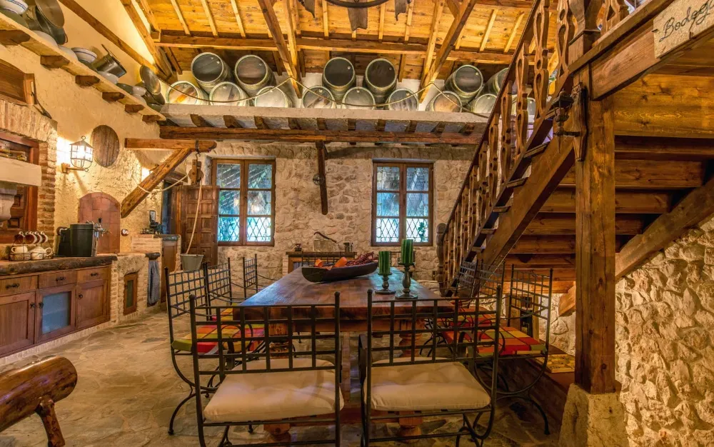 Comedor rústico con una larga mesa de madera, sillas de metal, paredes de piedra y barriles de vino almacenados en una viga del techo elevado.