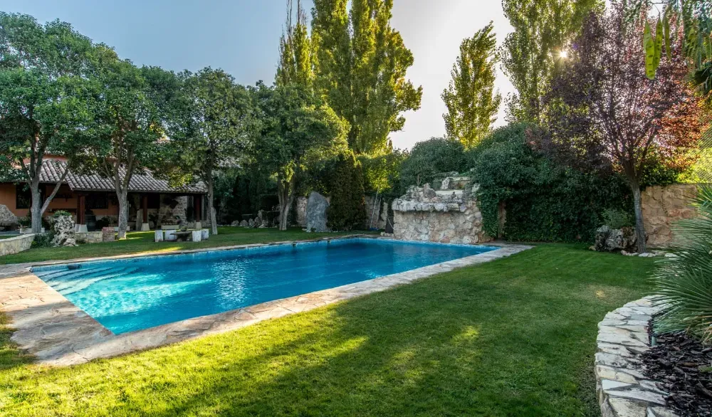 Una piscina rectangular rodeada de césped, árboles y muros de piedra en un día soleado.