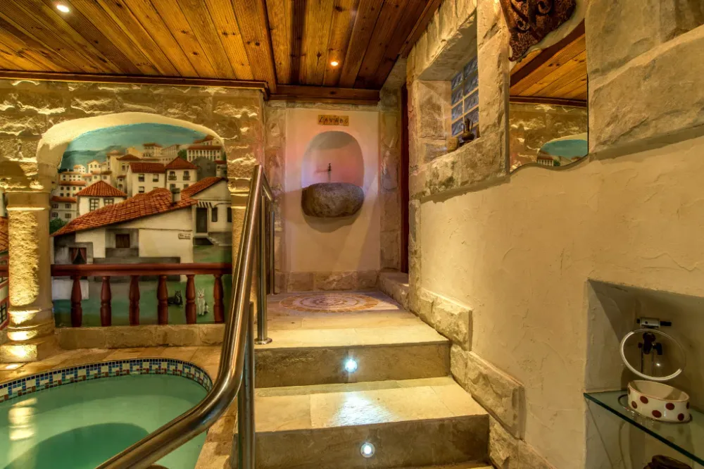 Zona de piscina cubierta con paredes de piedra, techo de madera y un mural de un pueblo costero, que incluye escaleras y una barandilla metálica.