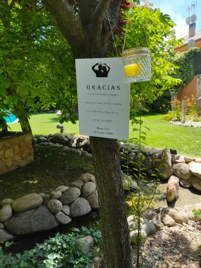 Un cartel blanco cuelga de un árbol en un jardín soleado, con un farol de velas al lado. El cartel dice 