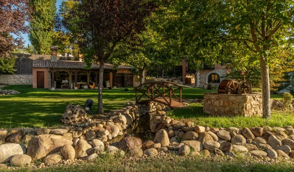 Un puente peatonal de madera cruza un pequeño arroyo en un jardín soleado y verde que cuenta con un pozo de piedra y una casa rústica.