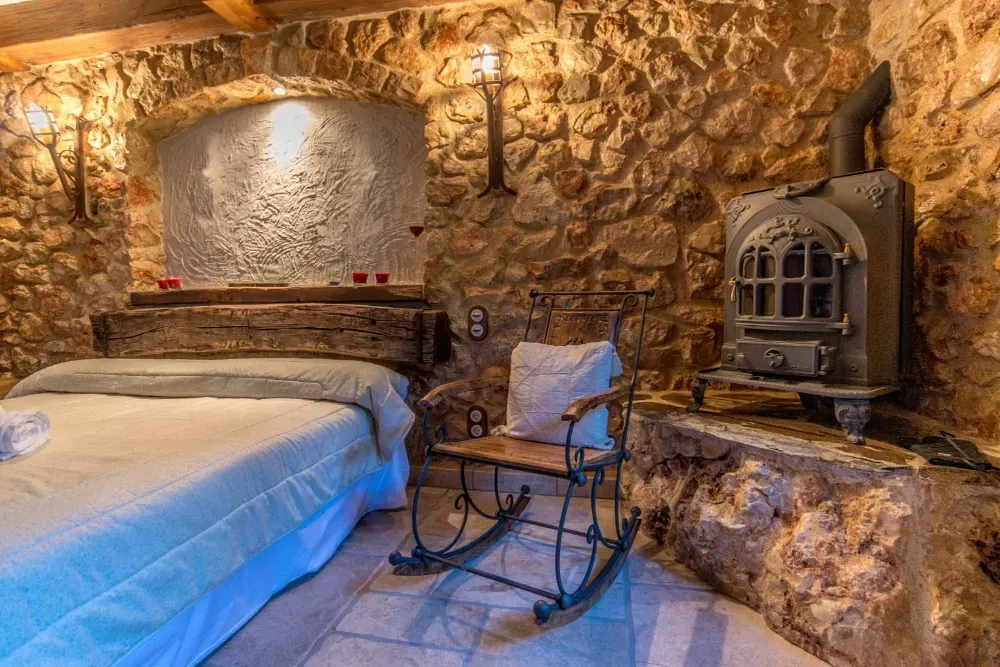 Un dormitorio rústico con paredes de piedra, una cama, una mecedora y una chimenea de hierro fundido.