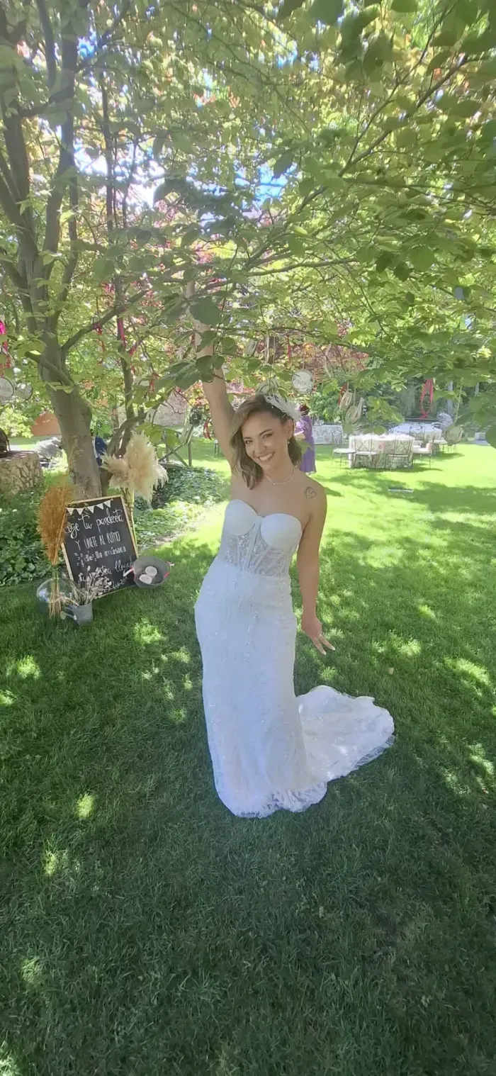 Una persona sonriente, vestida con un vestido de novia blanco sin tirantes, se encuentra de pie bajo un árbol en un jardín soleado y cubierto de césped.