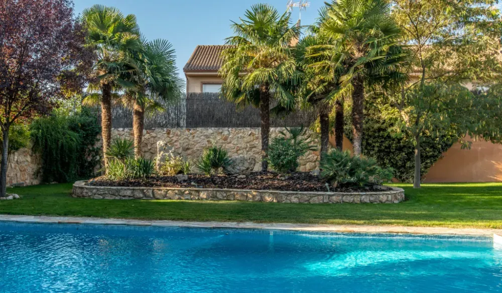 Una piscina azul en primer plano, con palmeras y un muro de contención de piedra en un jardín soleado al fondo.