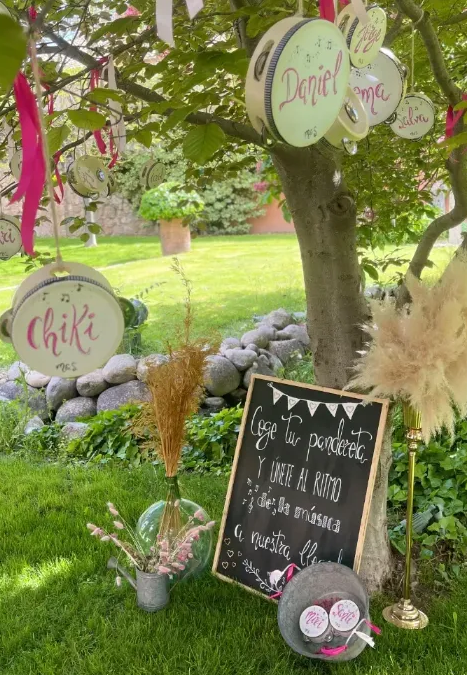 Una escena en el césped con panderetas colgando de un árbol, un cartel de pizarra y decoraciones de hierba de la Pampa para un evento al aire libre.