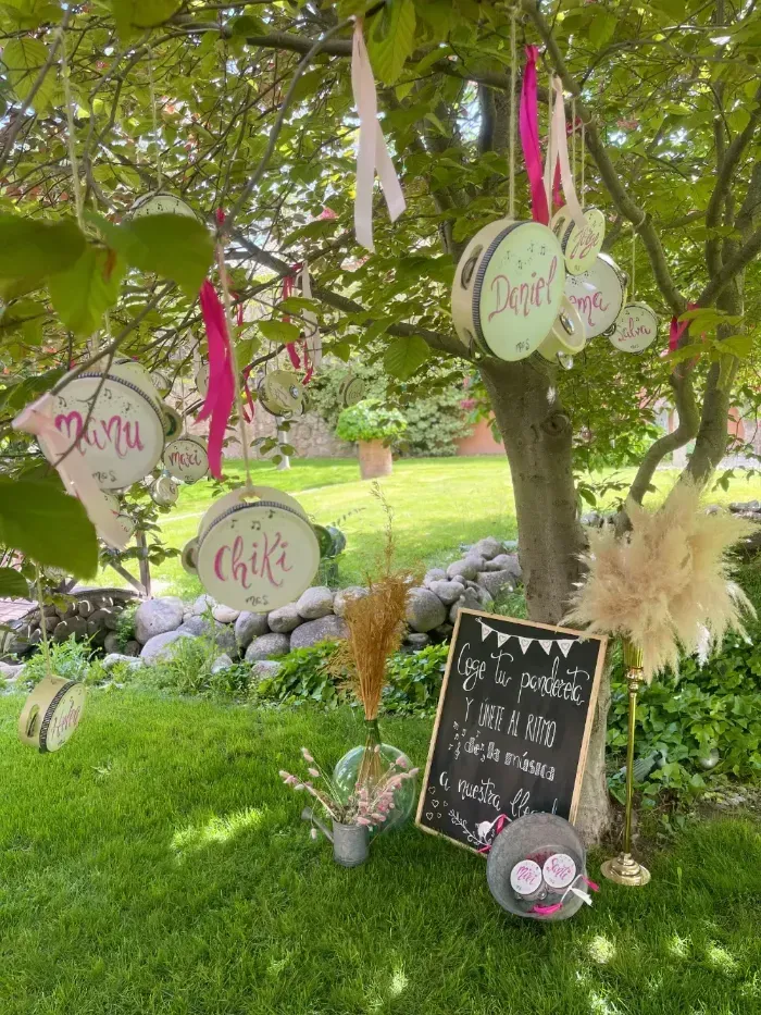 En un jardín cubierto de césped, cuelgan panderetas decoradas con nombres grabados de las ramas de los árboles, encima de una pizarra colocada sobre un caballete.