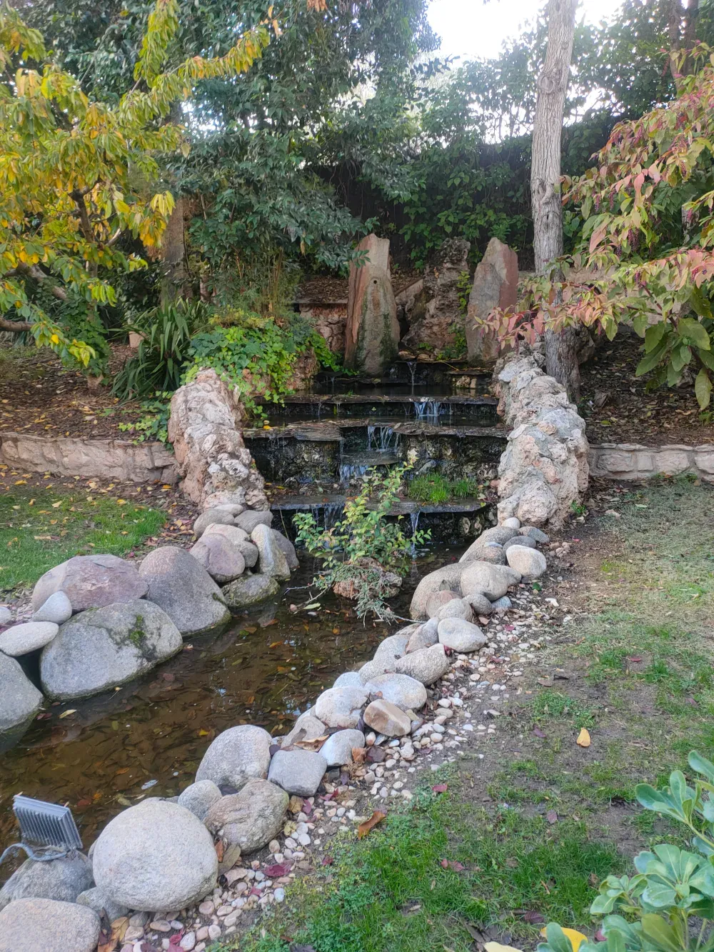 Una cascada escalonada que desemboca en un estanque poco profundo revestido de piedra y rodeado de árboles y vegetación.