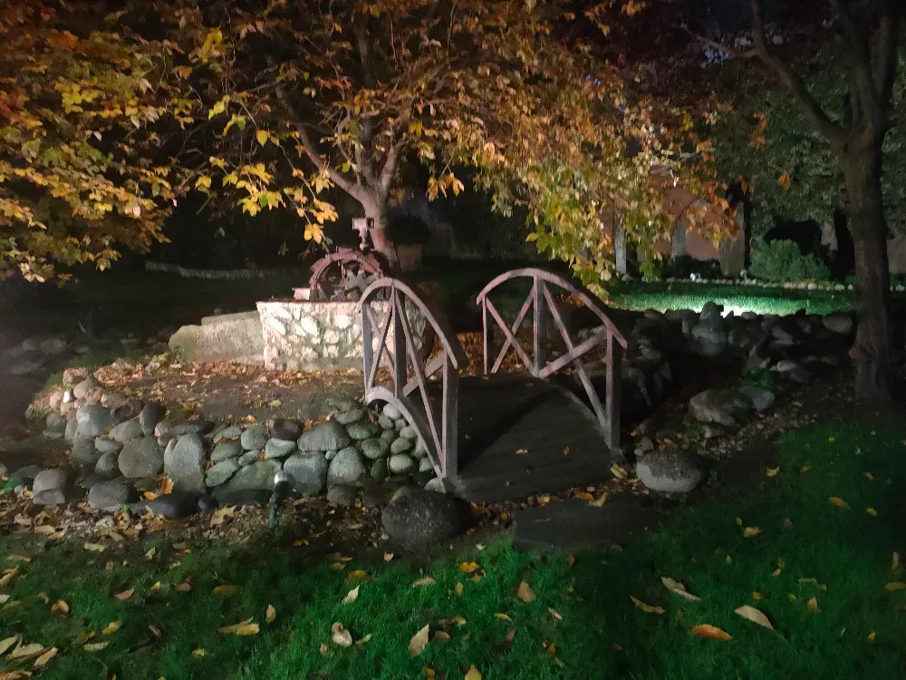 Un puente de piedra conduce a una pequeña zona ajardinada iluminada que cuenta con un muro de piedra, una noria y árboles con hojas otoñales.