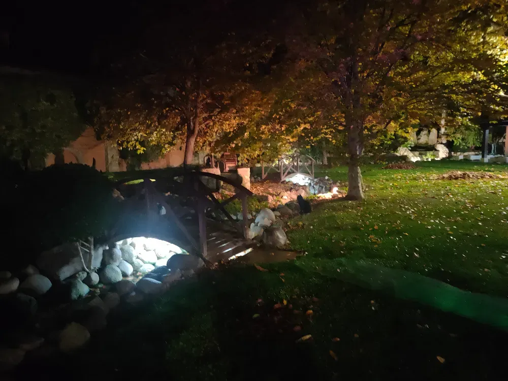 Por la noche, una pasarela de madera iluminada con luces brillantes cruza un pequeño arroyo de jardín, enclavada en un patio cubierto de césped y repleto de árboles.