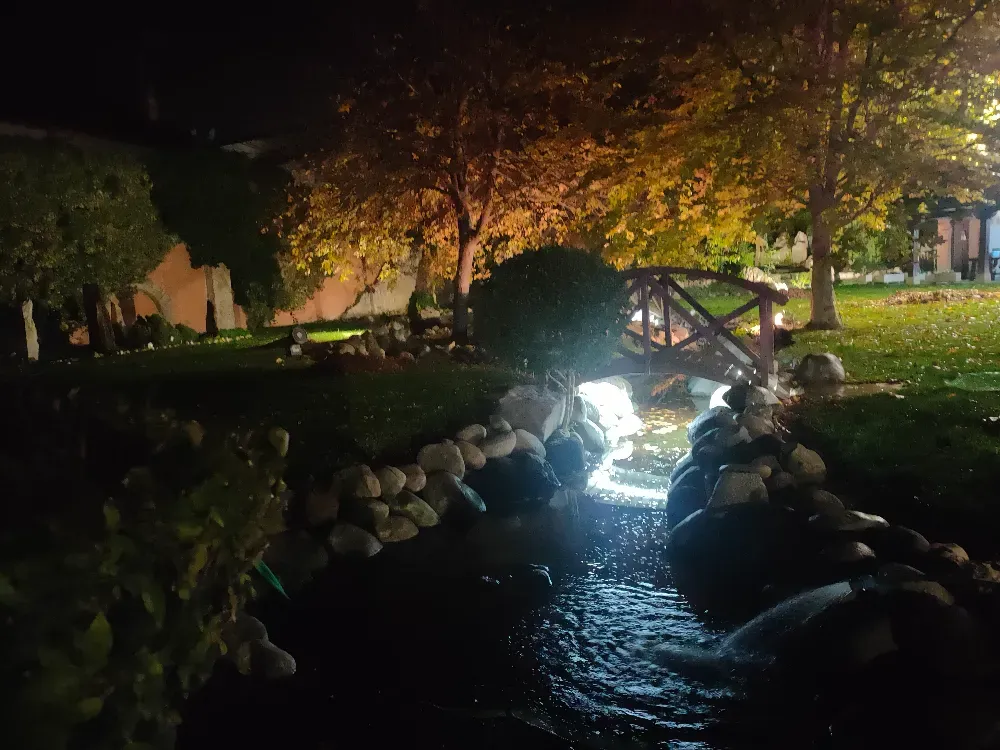 Un pequeño arroyo de piedras fluye bajo un puente peatonal de madera en un jardín iluminado por una cálida iluminación paisajística durante la noche.