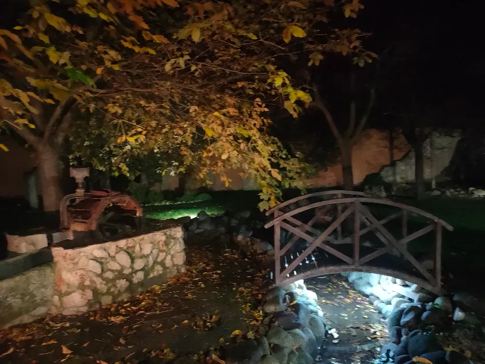 Una escena nocturna al aire libre iluminada, con un pequeño puente peatonal de madera sobre un arroyo, un muro de piedra y árboles otoñales.