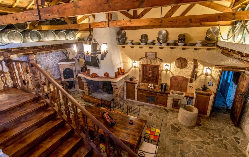 Habitación rústica con techos altos, escaleras de madera, una gran chimenea central, suelos de piedra y elementos decorativos en las paredes.