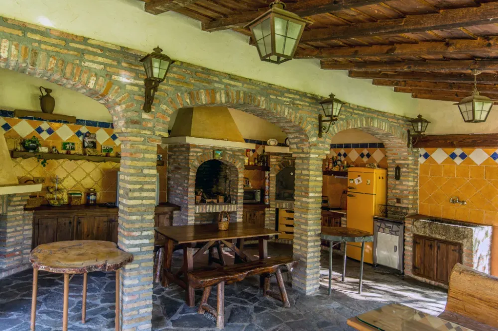 Una cocina rústica al aire libre, construida en ladrillo, con suelos de piedra, techo con vigas de madera, faroles colgantes y un frigorífico amarillo.