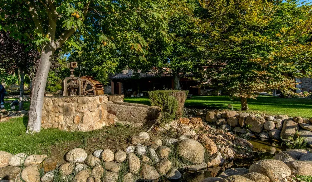 Un pozo de piedra con un antiguo mecanismo de poleas se encuentra junto a un arroyo rocoso en un jardín exuberante y lleno de árboles, en un día soleado.