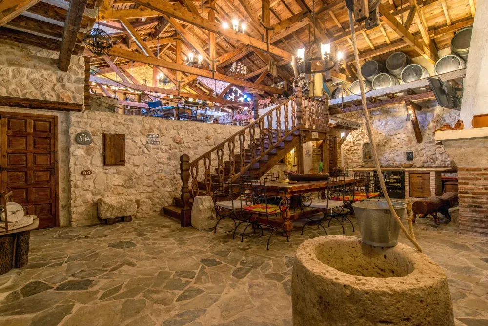 Una habitación rústica interior con paredes de piedra, una escalera de madera, vigas a la vista, una mesa central y un pozo decorativo de piedra.