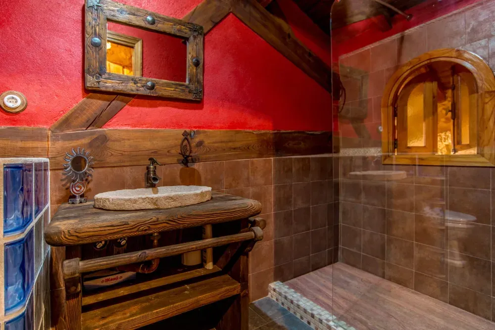 Baño rústico con lavabo de piedra, tocador de madera, paredes de color rojo intenso y zona de ducha alicatada con ventana arqueada.