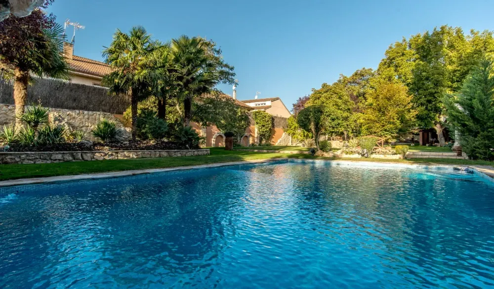Una piscina rectangular de agua azul brillante se encuentra en un jardín exuberante con árboles verdes y un muro de piedra como telón de fondo.