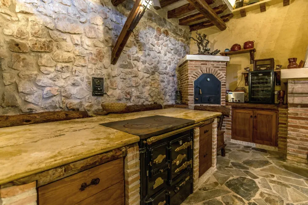 Una cocina rústica con paredes de piedra, vigas de madera, una gran estufa negra de estilo vintage, encimeras de granito y baldosas de piedra en el suelo.