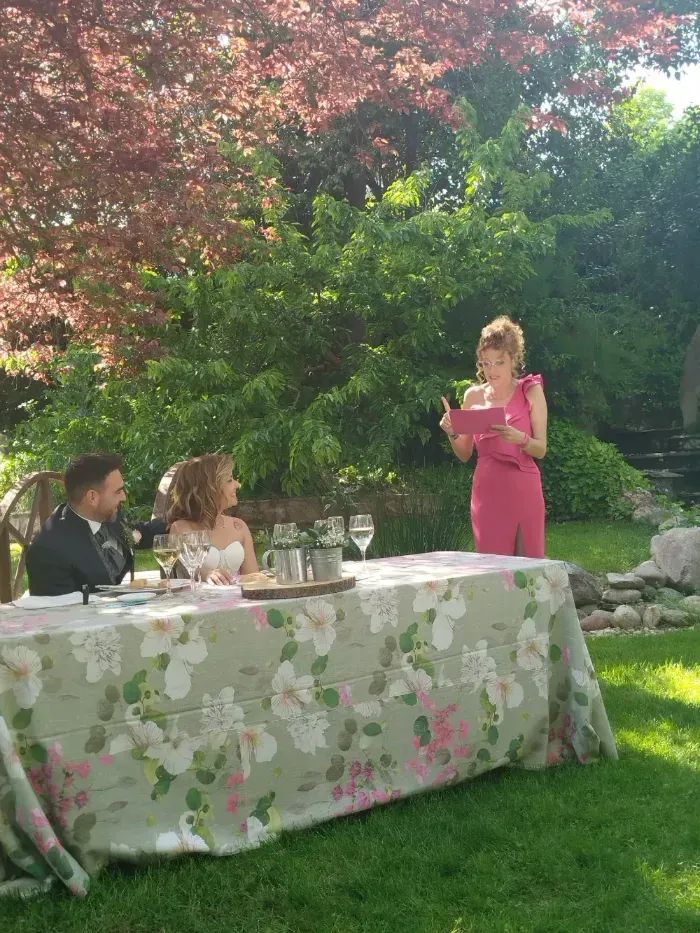 Una persona vestida con un vestido rosa lee unas notas en una boda en un jardín, mientras una pareja se sienta cerca en una mesa cubierta de flores.