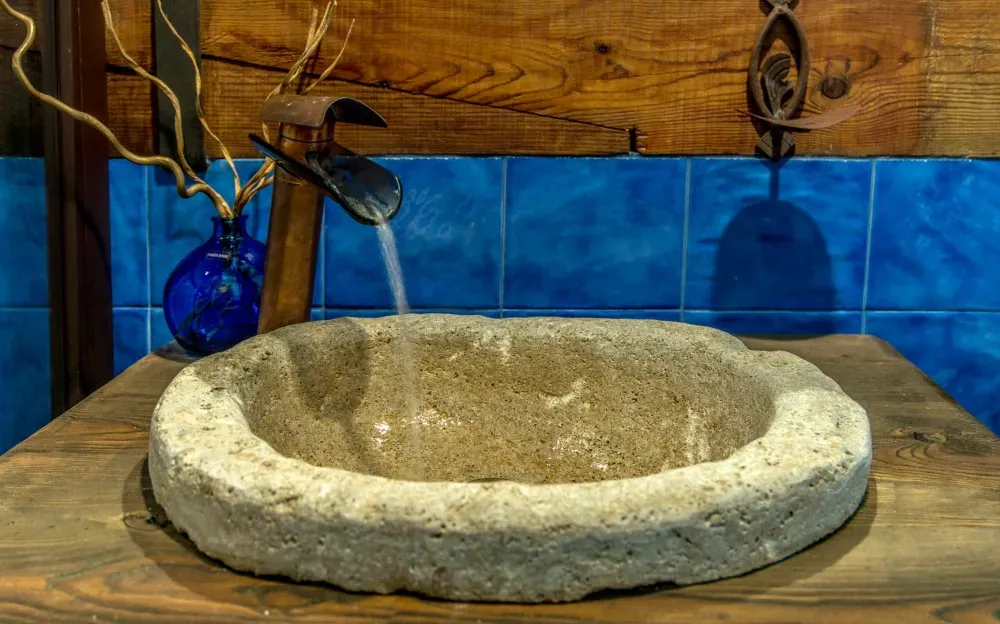 Un lavabo de piedra descansa sobre una encimera de madera con un grifo metálico moderno que dispensa un chorro de agua sobre azulejos azules.