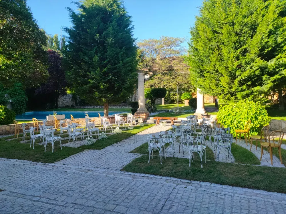 Filas de sillas blancas dispuestas para una ceremonia al aire libre en un patio de piedra y césped, con árboles y una piscina en la parte trasera.