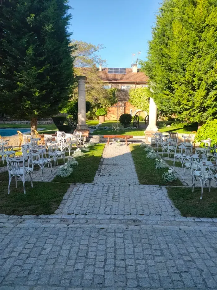 Un montaje para ceremonia de boda al aire libre con filas de sillas blancas frente a un camino empedrado entre dos pilares de piedra.