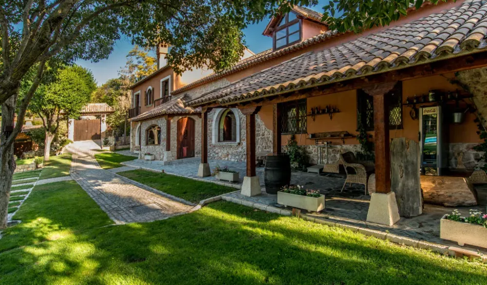 Una villa rústica de color terracota con detalles de piedra, techo de tejas y un patio de piedra sombreado con vistas a un césped verde.