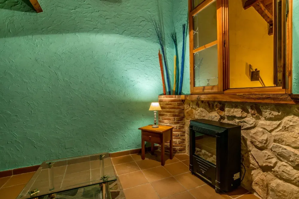 Una pared texturizada de color verde azulado sobre una base de piedra, con una pequeña mesa de madera con una lámpara, un calefactor y una mesa de cristal.