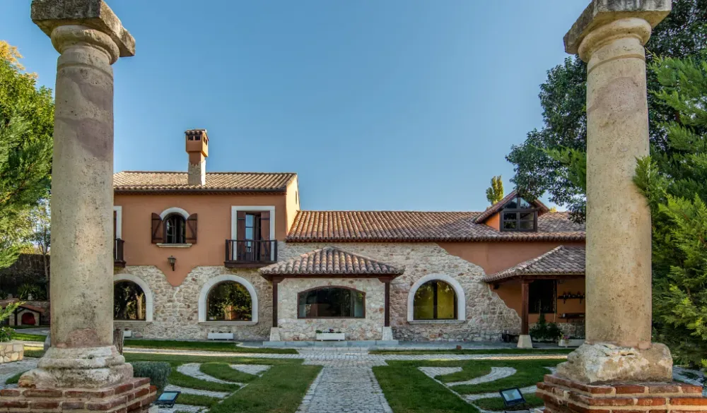 Una villa rústica con paredes de color tostado y detalles de piedra detrás de dos pilares de piedra, situada en un jardín con un sendero pavimentado.