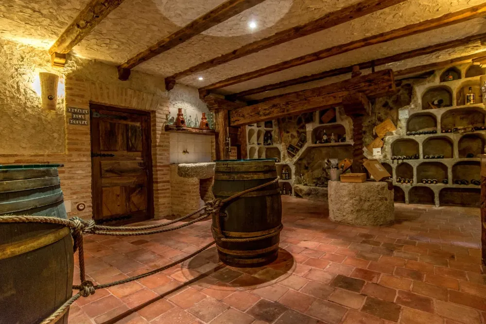 Una bodega rústica con paredes de piedra, vigas de madera en el techo, suelos de terracota, barricas de vino y nichos abovedados.