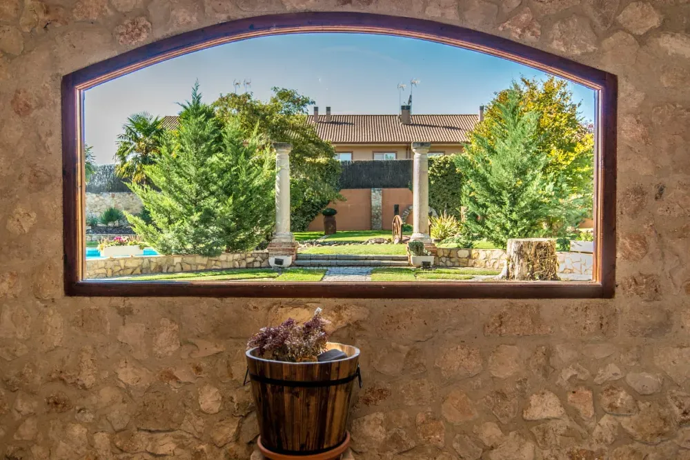 Un muro de piedra enmarca un gran ventanal arqueado con vistas a un jardín cubierto de césped, con dos columnas de piedra y grandes árboles verdes.