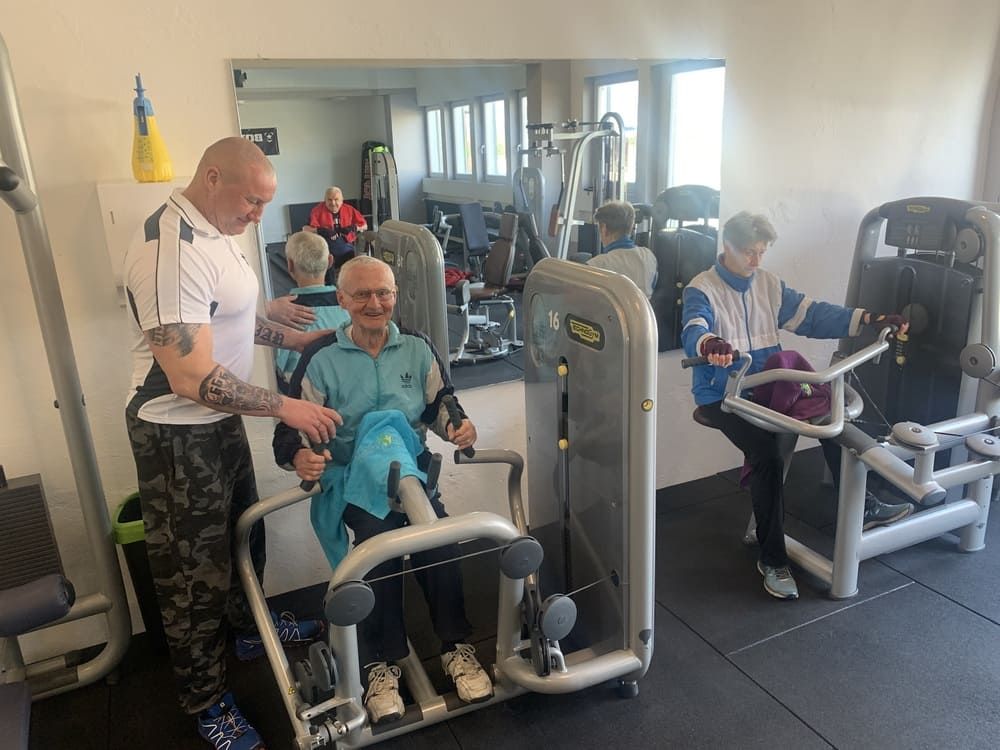 Ein Mann hilft einem älteren Mann auf einer Maschine in einem Fitnessstudio | bodyfit24