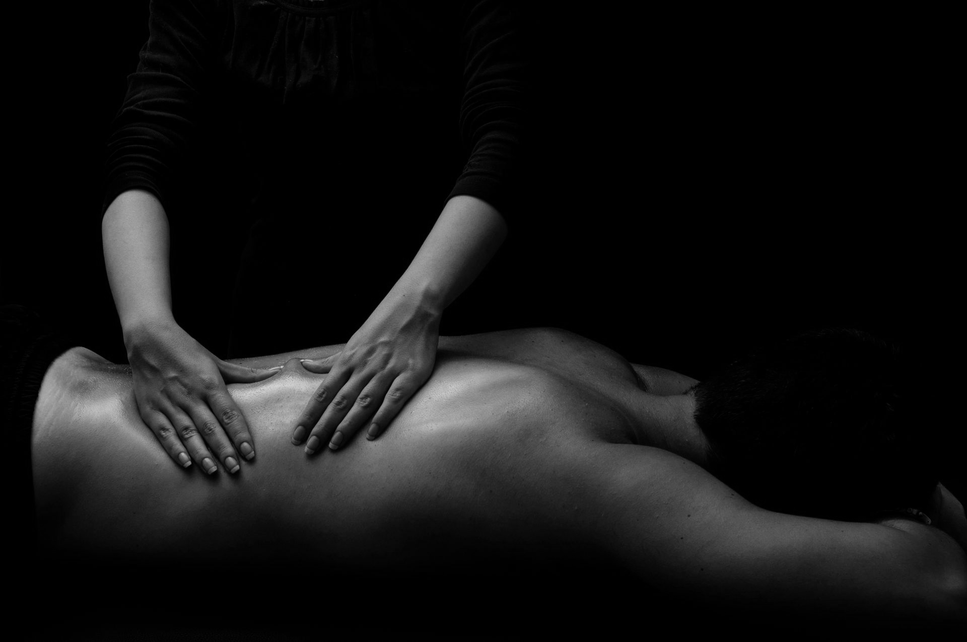 Une personne reçoit un massage du dos dans un environnement faiblement éclairé, présenté en noir et blanc à fort contraste.