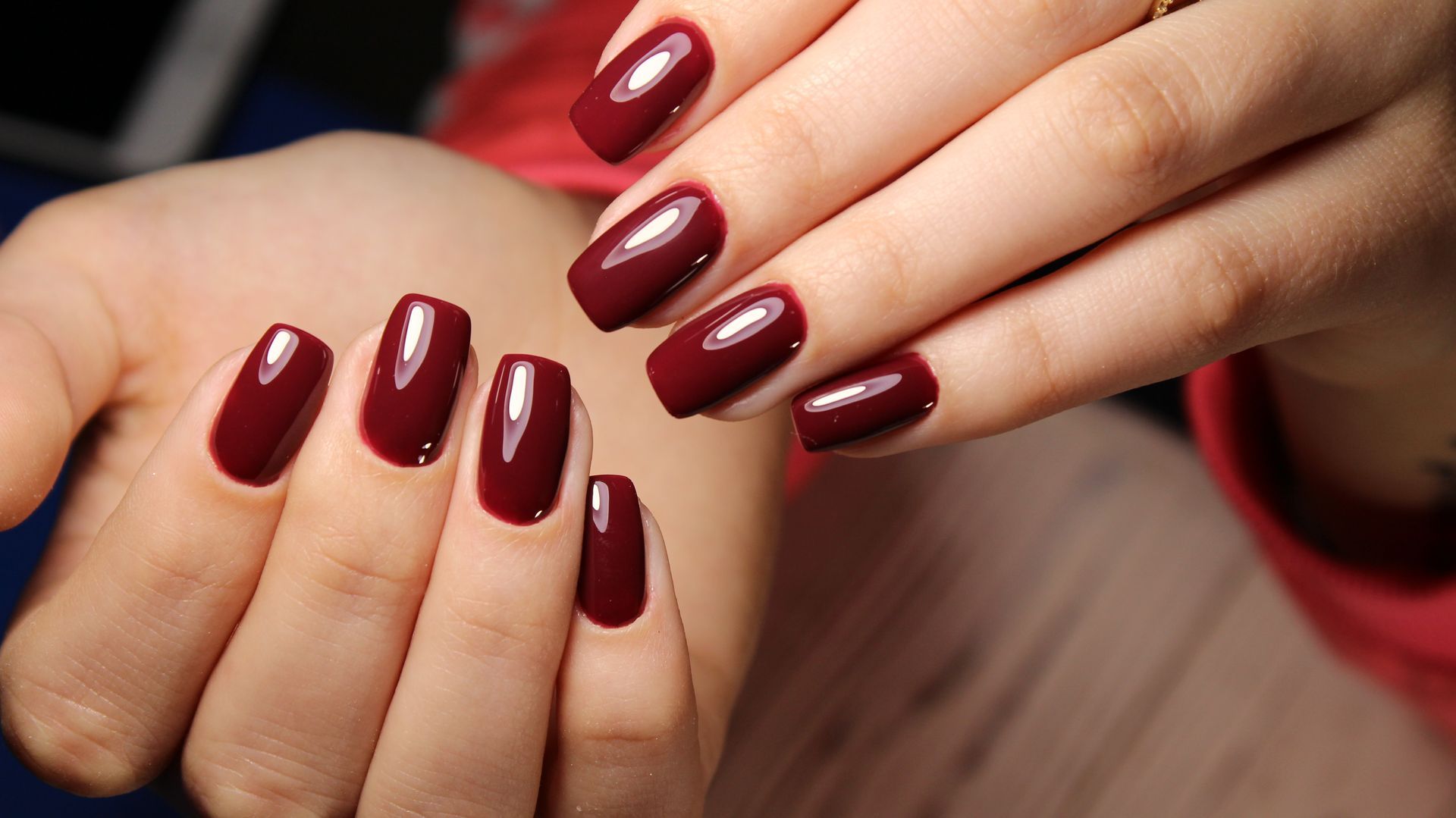 Deux mains arborant une manucure professionnelle avec des ongles carrés, brillants, de couleur bordeaux foncé.