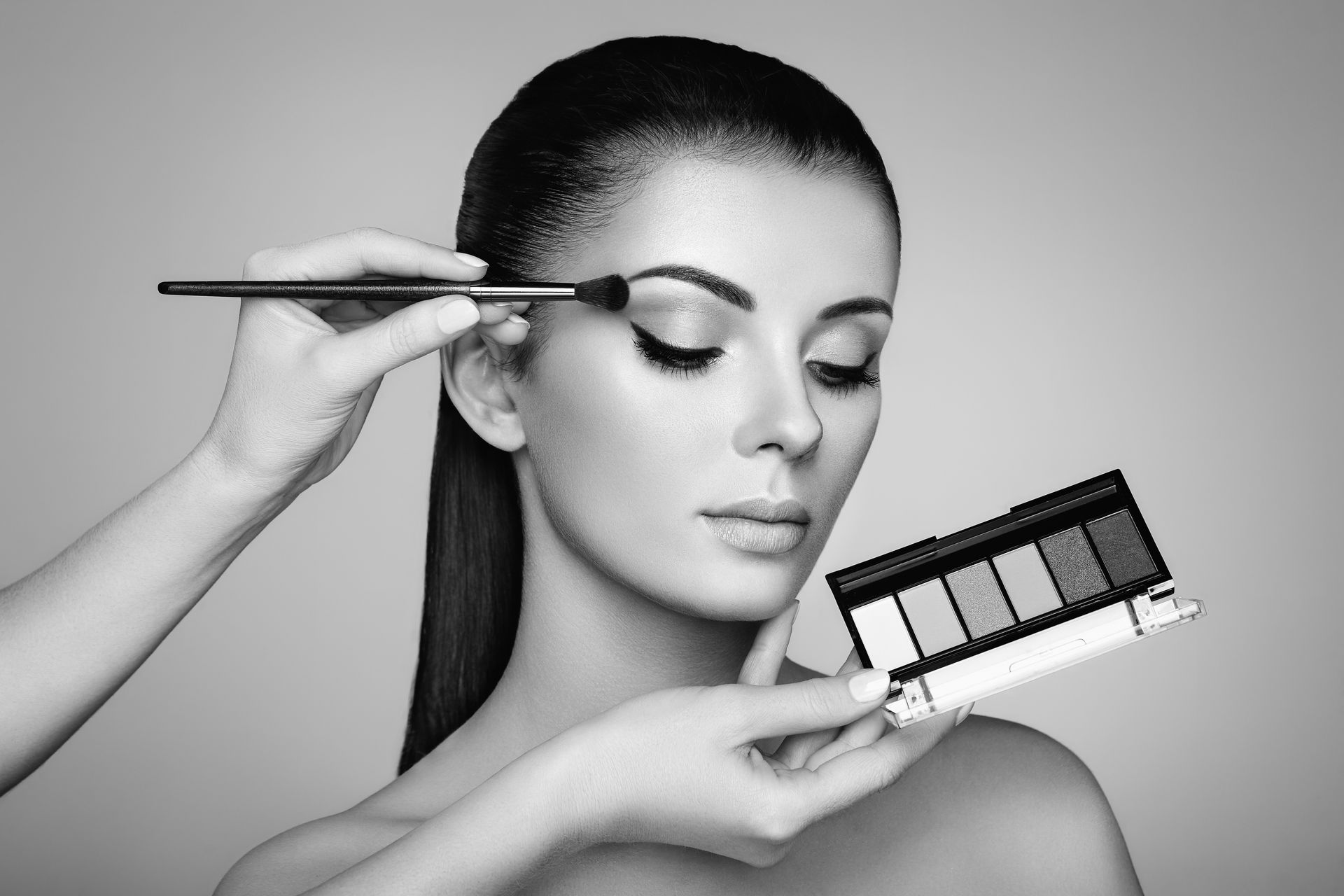 On applique du maquillage sur les yeux d'une personne à l'aide d'un pinceau, tout en tenant une palette de fards à paupières noirs et blancs.