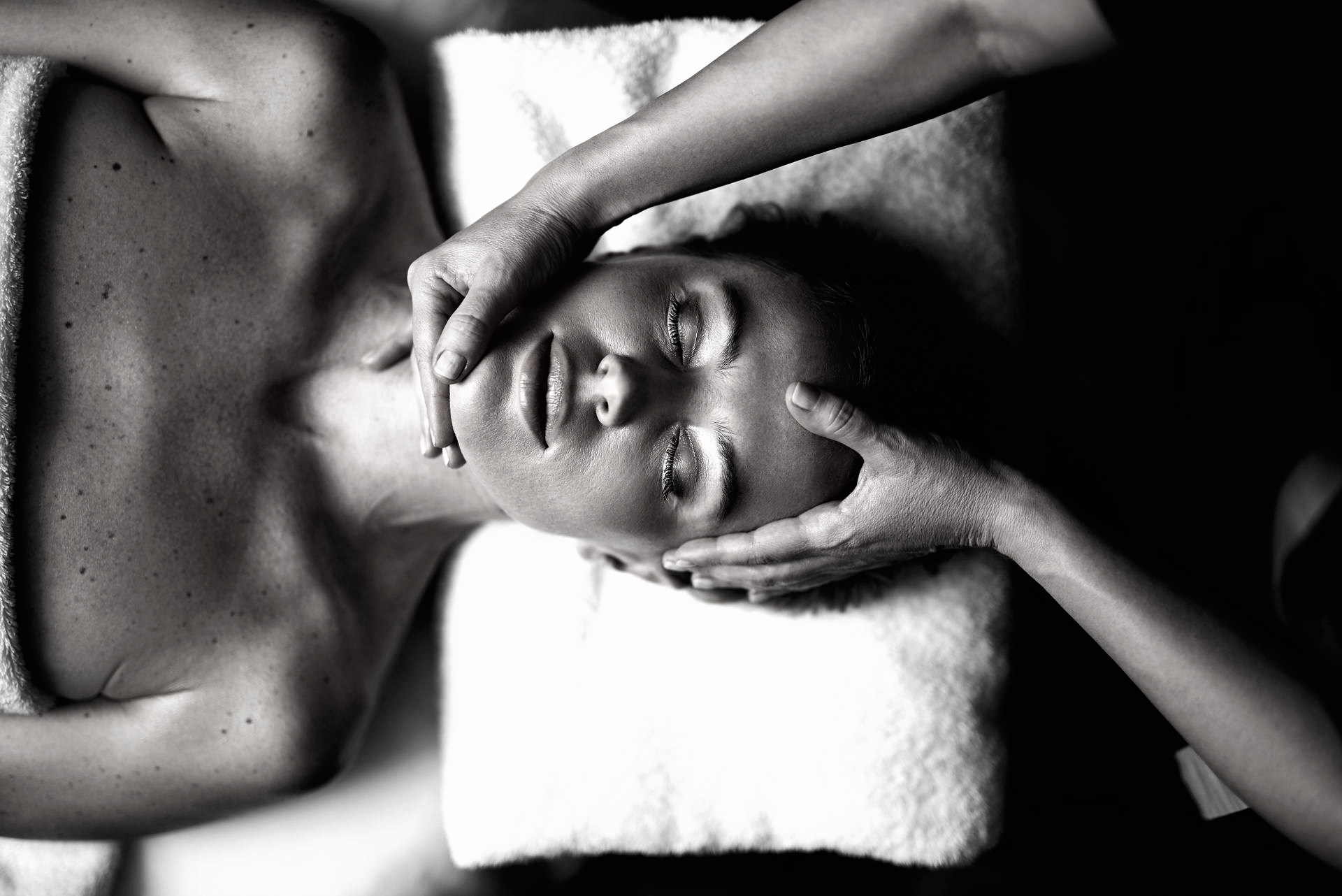 Vue en noir et blanc d'une personne recevant un massage facial relaxant, allongée sur une surface rembourrée.