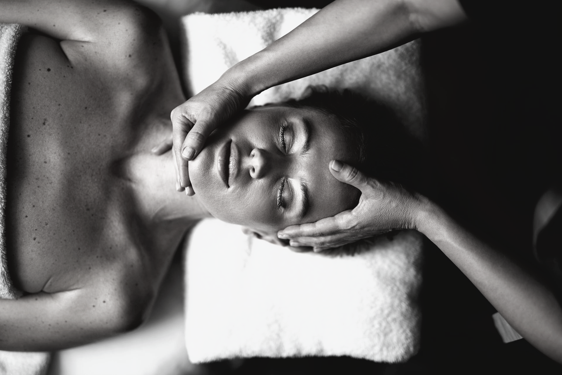Gros plan en noir et blanc d'une personne recevant un massage facial relaxant, allongée sur une surface blanche et douce.