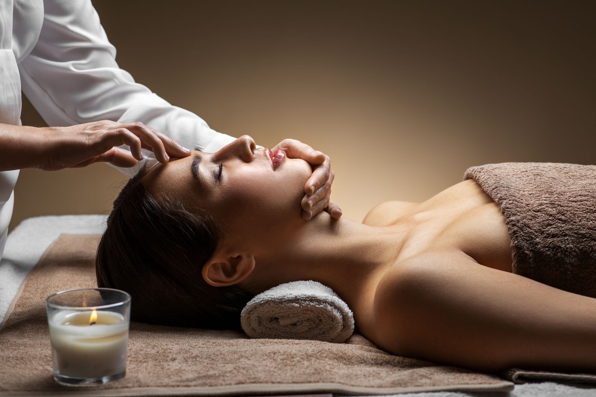 Une personne allongée sur une table de spa reçoit un massage facial relaxant, dans une ambiance tamisée et calme, à la lueur d'une bougie allumée.