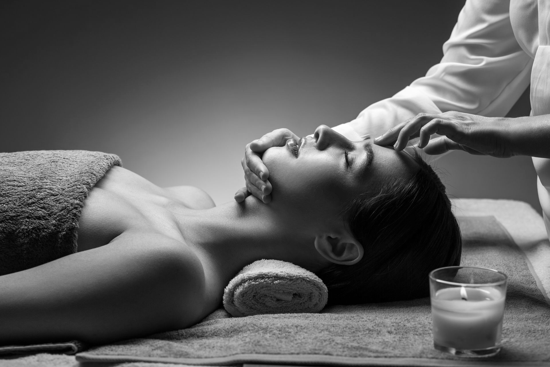 Photo en noir et blanc d'une personne recevant un massage facial professionnel, avec une bougie allumée sur la table à proximité.