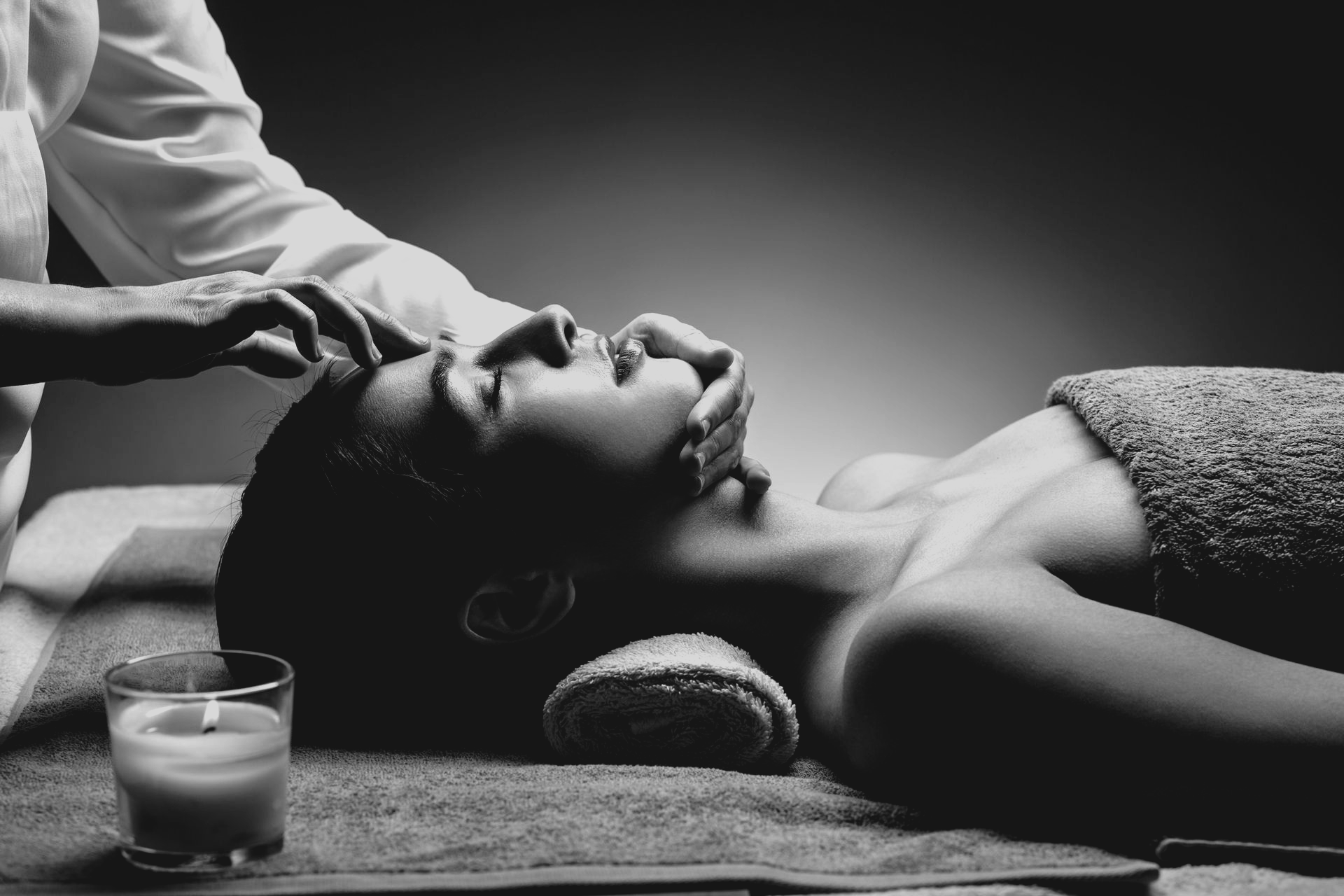 Une personne reçoit un massage facial relaxant dans un spa à l'atmosphère sereine, avec une bougie au premier plan. Noir et blanc.